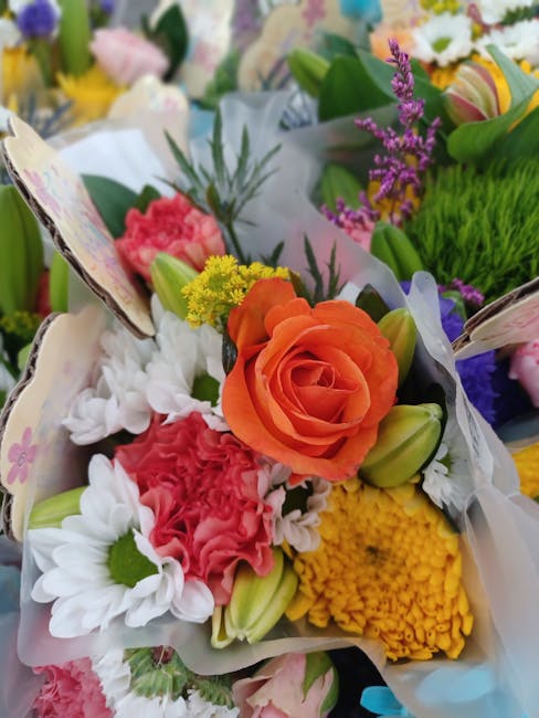 A vibrant floral bouquet featuring orange roses, white daisies with green centers, pink carnations, yellow chrysanthemums, and purple lavender accents, wrapped in white floral paper. The arrangement i