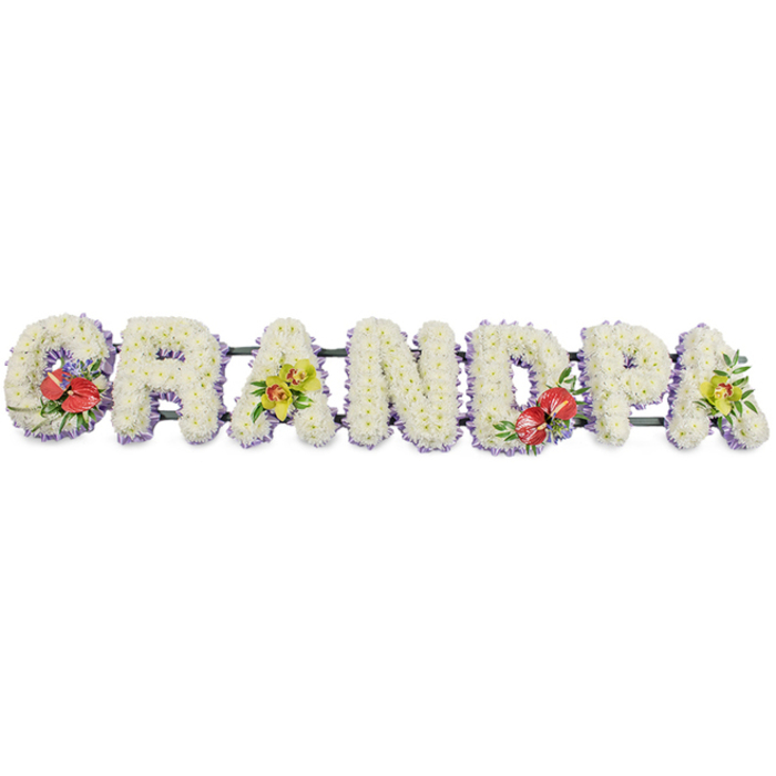 The image shows a carefully crafted funeral flower tribute spelling "GRANDPA" in proud, block capital letters, arranged in a straight horizontal line against a plain white background so the design can be seen clearly from a distance. Each letter is fully packed with small white chrysanthemum blooms, placed closely together so that no gaps are visible, creating a smooth, pillow-like texture that symbolises purity and gentle farewell. At the base of each letter, a soft band of lavender ribbon peeps through, encircling the framework and giving a cool, light purple edge that subtly defines every curve and corner without overpowering the calm white. Dark green metal or wooden bars run along the back of the word, joining the letters into one continuous tribute that can be easily positioned at a funeral in Stanmore, perhaps in a local chapel near Belmont Circle or within the peaceful grounds around Canons Park. Splashes of vivid colour bring a deeply personal touch: the "G" on the far left and the "D" near the centre carry matching sprays of deep red anthuriums, their glossy, heart-shaped spathes and yellow-tipped spadices standing proudly among lush foliage and hints of smaller accent flowers. The first and final "A" are dressed with clusters of yellow-green orchids, likely cymbidiums, their elegant, arching petals and speckled throats lending an air of refinement and enduring beauty. Together, these elements form a bespoke GRANDPA name tribute, a piece that families might order when they want to honour a grandfather's memory with something both traditional and distinctive, conveying love, respect, and gratitude at a Stanmore funeral or graveside gathering.